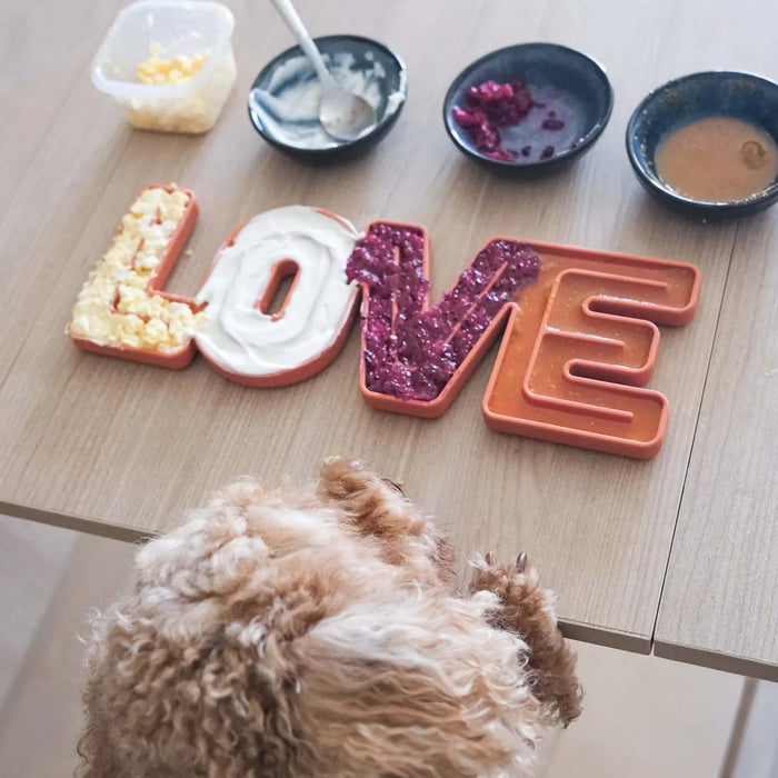 Dog perched against a table looking at lick mat reading love with small bowls filled with food
