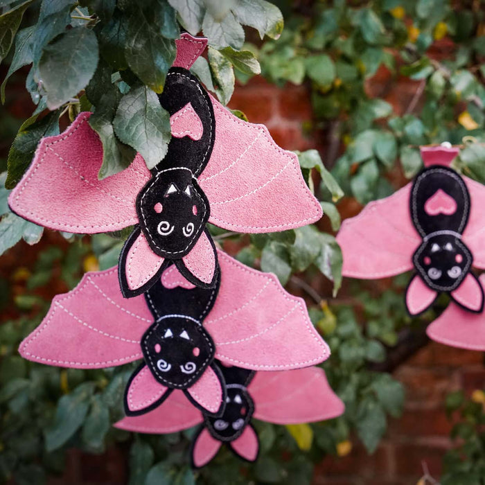 Pink and black felt bat decorations hanging from green tree branches outdoors, with a blurred brick wall and foliage in the background.