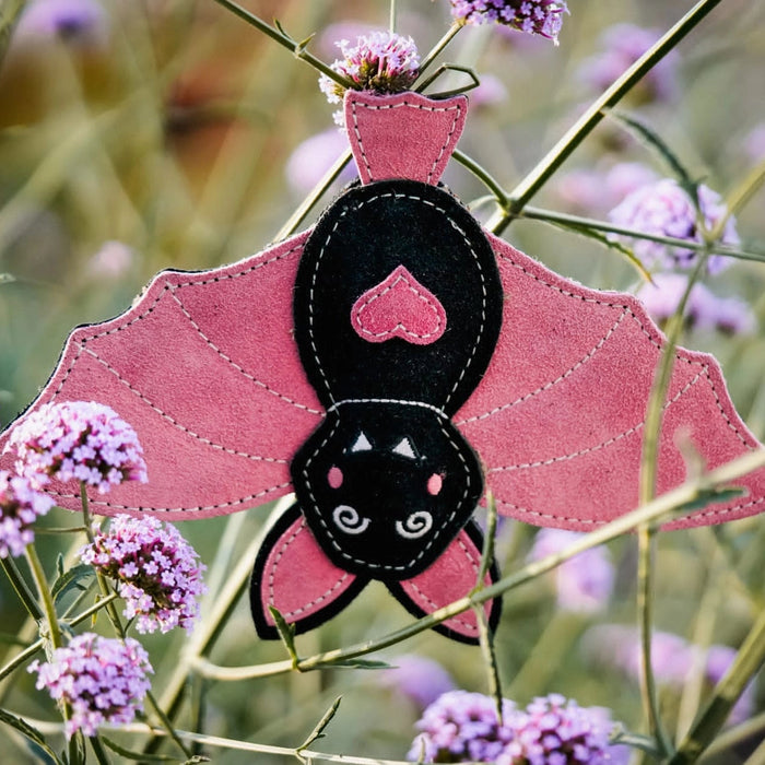 A handmade pink and black felt bat ornament hanging among purple flowers and green stems in an outdoor garden setting.
