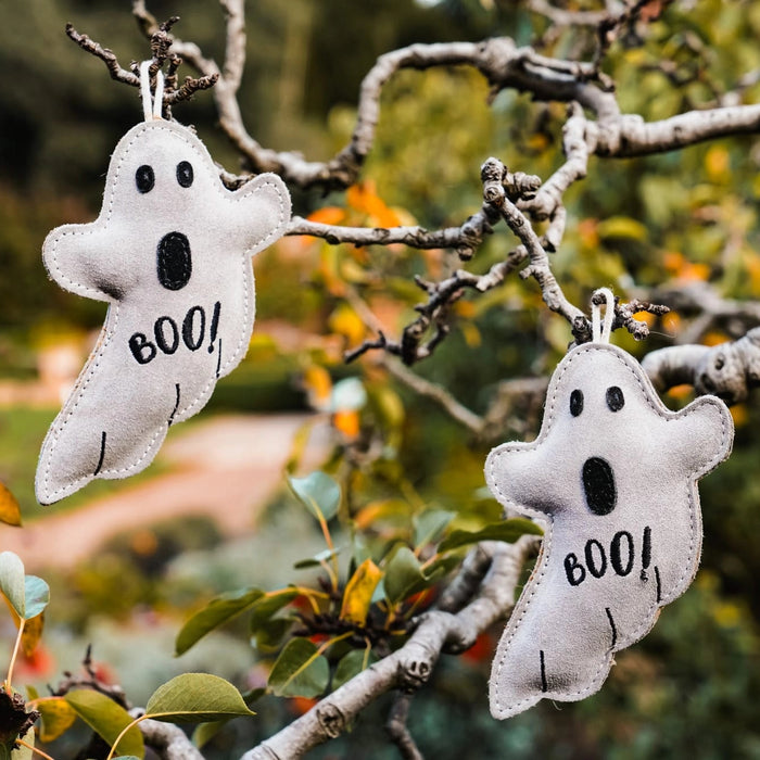 Two white felt ghost ornaments with black embroidered eyes, mouths, and the word 'BOO!' hanging from tree branches outdoors.