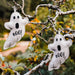 Two white felt ghost ornaments with black embroidered eyes, mouths, and the word 'BOO!' hanging from tree branches outdoors.