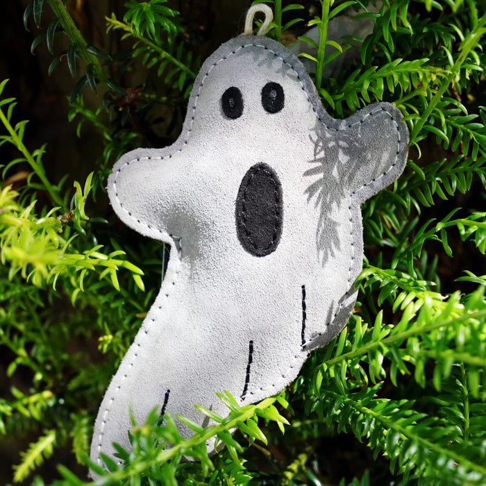 A small ghost-shaped felt ornament or decoration in light gray with black embroidered facial features and stitched details, hanging among bright green pine branches.