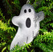 A small ghost-shaped felt ornament or decoration in light gray with black embroidered facial features and stitched details, hanging among bright green pine branches.