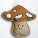 A handmade felt mushroom-shaped hanging ornament or pouch, featuring a brown cap with white spots, a beige stem with a simple embroidered smiley face, and a white hanging loop at the top, shown against a plain white background.