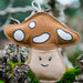 A handmade felt mushroom ornament with a brown cap, white spots, and a beige stem with a simple embroidered smiley face, hanging from a loop on a mossy branch.