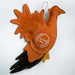 A handmade felt Christmas ornament shaped like a turkey, featuring a warm orange-brown body and tail, a black beak, a small mustard yellow wing, and white contrast stitching. A circular white logo patch reading 'the orange farm' is stitched onto the side, and a white hanging loop is attached at the top.