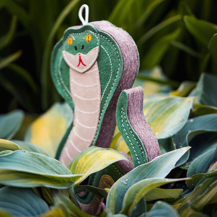 A felt snake garden ornament with a green head and body, a purple tail, and a beige underbelly, nestled among green and yellow hosta leaves.