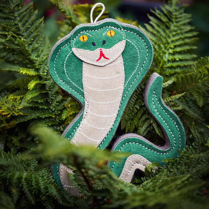 A green and beige felt snake ornament with embroidered details, nestled among green fern leaves.