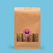 A kraft paper stand-up pouch with a clear window showing several brown rolled sticks or bars inside, centered on a solid light blue background. A pink circular sticker with a dark blue 'w' logo is on the front.