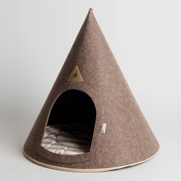 A cone-shaped felt pet house in a warm taupe-brown color, featuring a dark arched entrance with a striped cushion inside and a small triangular metal brand plaque on the front.