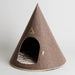 A cone-shaped felt pet house in a warm taupe-brown color, featuring a dark arched entrance with a striped cushion inside and a small triangular metal brand plaque on the front.
