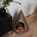 A small brown poodle lies inside a cone-shaped felt pet house with a rounded base, placed on a light wood floor next to a black cabinet with a potted green plant.