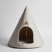 A cone-shaped pet house made of light gray felt, featuring a triangular metal brand plaque on the front and a striped cushion inside, photographed on a neutral light gray background.