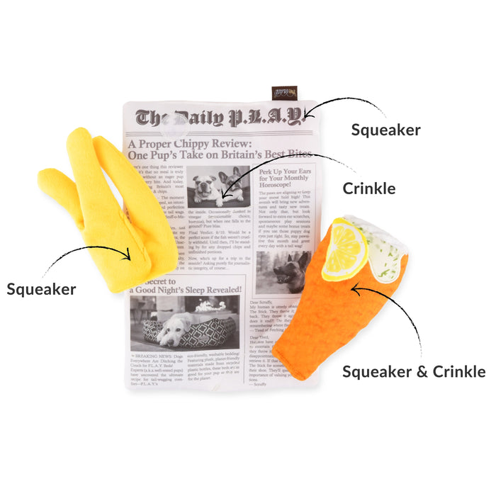 Two novelty dog toys shaped like gloves are displayed on a newspaper prop against a white background, with black callout lines and text labels identifying the yellow toy as Squeaker and the orange toy as Squeaker & Crinkle.