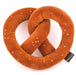 A plush pretzel-shaped pillow in a warm brownish-orange color, decorated with small white faux-seed details and a small black brand tag on the right side, shown on a plain white background.