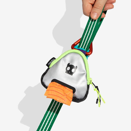 A hand holds a green webbed pet leash with a mushroom-shaped metal tag. The tag has a silver reflective body, a black cartoon face, an orange faux-fur detail, and a red carabiner, shown against a light gray background.