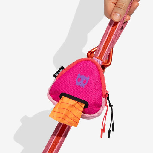 A small pink pet poop bag with a purple cartoon face logo, attached to a pink and orange striped leash. A hand is holding the leash at the top, and the bag is shown dispensing a small orange poop training mat against a light gray background.