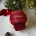 A plush red and cream knit sweater dog toy shaped like a cupcake, with 'PUPS & BUBS' embroidered in gold on the red frosting. The toy is styled on a white textured surface with Christmas decorations, including pine branches, a red ornament, and a pinecone.
