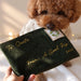 A brown poodle holds a dark green velvet Christmas letter envelope with gold foil writing and a decorative stamp, while a hand holds the bottom corner. The background is softly blurred with warm bokeh lights.