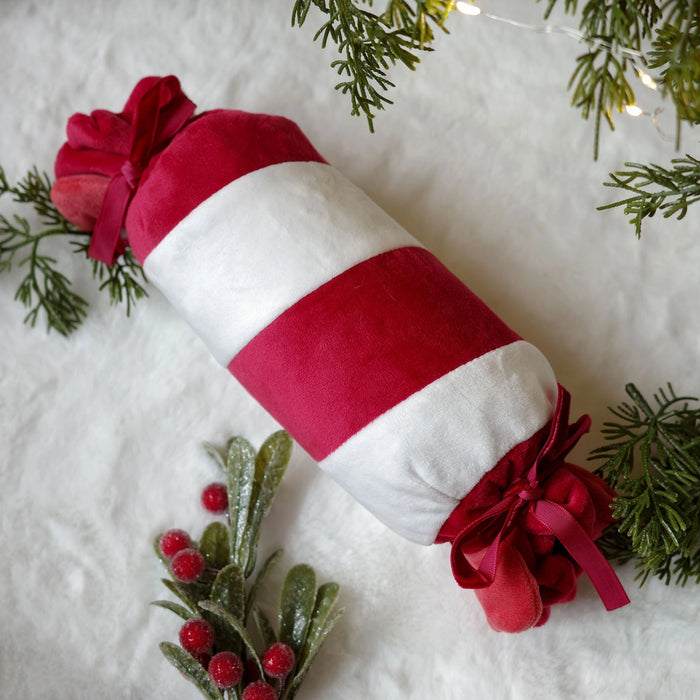 A cylindrical Christmas ornament or decorative pillow with alternating red and white stripes, tied with red ribbon bows at both ends, displayed on a white textured surface with festive green pine branches and small red berries.