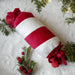 A cylindrical Christmas ornament or decorative pillow with alternating red and white stripes, tied with red ribbon bows at both ends, displayed on a white textured surface with festive green pine branches and small red berries.