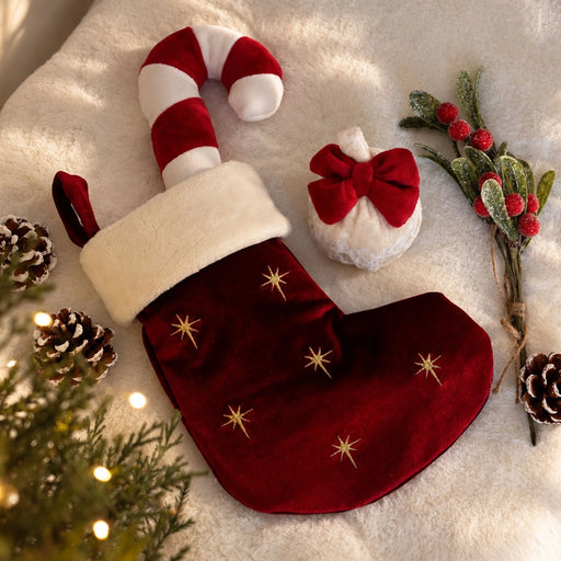 A red velvet Christmas stocking with gold star embroidery, a white faux-fur cuff, and a candy cane-shaped pull, styled on a white faux-fur surface with pinecones, evergreen branches, and a small white ornament with a red bow.