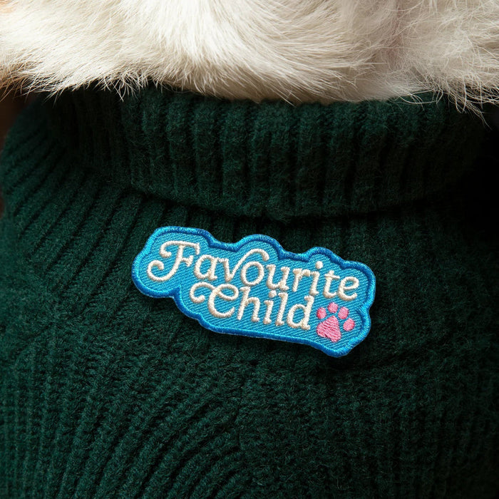 A close-up of a dark green ribbed knit pet sweater with a fluffy white faux-fur collar and a blue embroidered patch that reads 'Favourite Child' with a pink paw print.