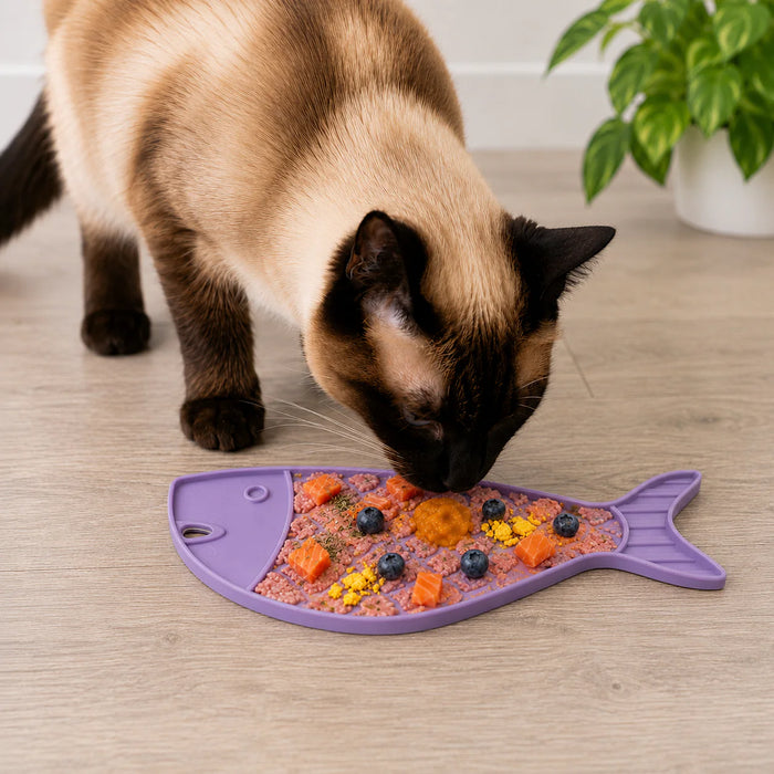 Cat eating from a fish-shaped purple mat with food on a wooden floor.