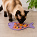 Cat eating from a fish-shaped purple mat with food on a wooden floor.