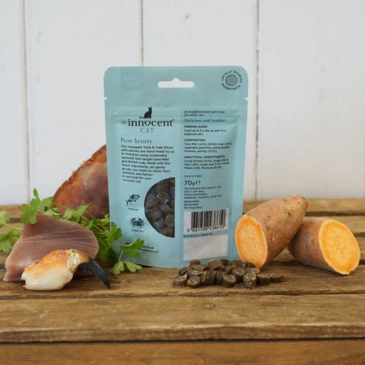 A stand-up pouch of Innocent Cat Pure Luxury wet cat food stands on a rustic wooden table, surrounded by food props including salmon, sweet potato, parsley, and a few kibble pieces. The pouch is light blue with black text and a clear window showing the wet food inside.