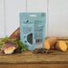 A stand-up pouch of Innocent Cat Pure Luxury wet cat food stands on a rustic wooden table, surrounded by food props including salmon, sweet potato, parsley, and a few kibble pieces. The pouch is light blue with black text and a clear window showing the wet food inside.