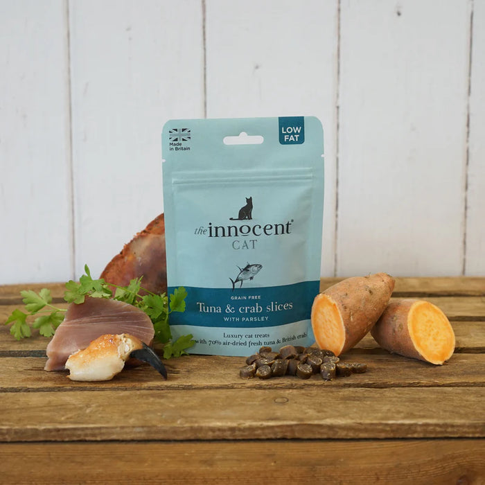 A package of The Innocent Cat adult-free tuna and crab slices for cats, styled on a rustic wooden table with raw ingredients (crab, parsley, sweet potato) and a few kibble pieces in front of it.