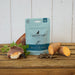 A package of The Innocent Cat adult-free tuna and crab slices for cats, styled on a rustic wooden table with raw ingredients (crab, parsley, sweet potato) and a few kibble pieces in front of it.