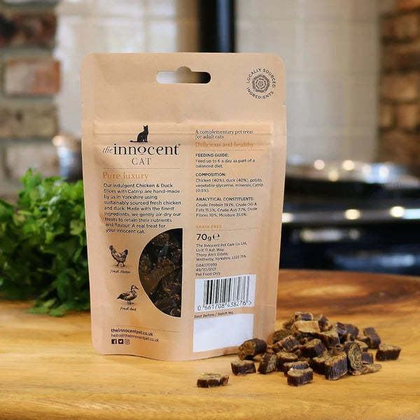 A 70g pouch of The Innocent Cat Pure Luxurys cat treats stands on a wooden surface with several dark brown chew pieces scattered in front. The background is a softly blurred kitchen setting.