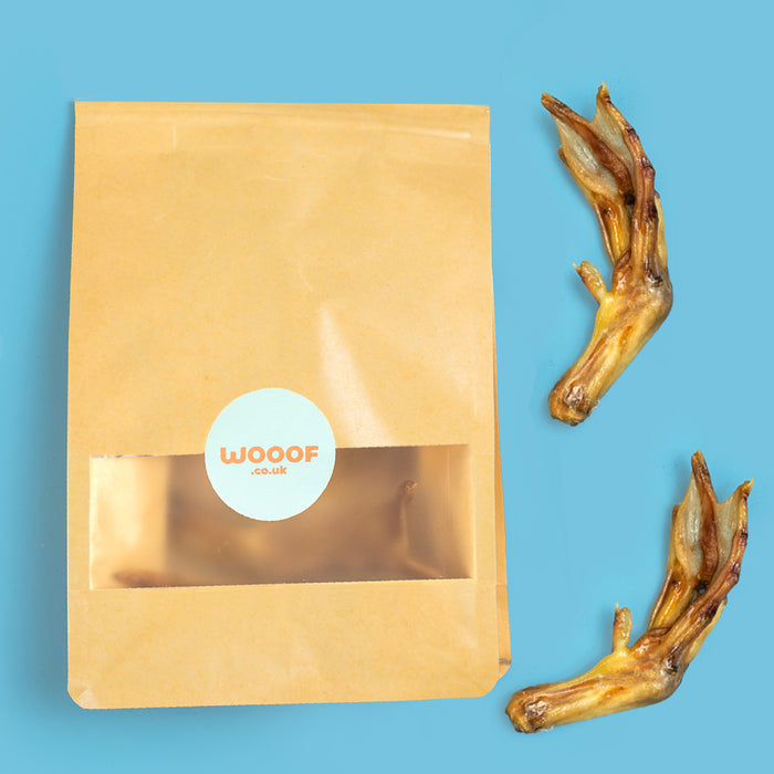 A paper bag with a clear window and a round white label reading 'WOOOF' sits on a bright blue background, alongside two dried chicken feet.