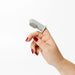 A hand with dark red painted nails holds a small light gray silicone nail care tool with a textured bristle head against a plain white background.