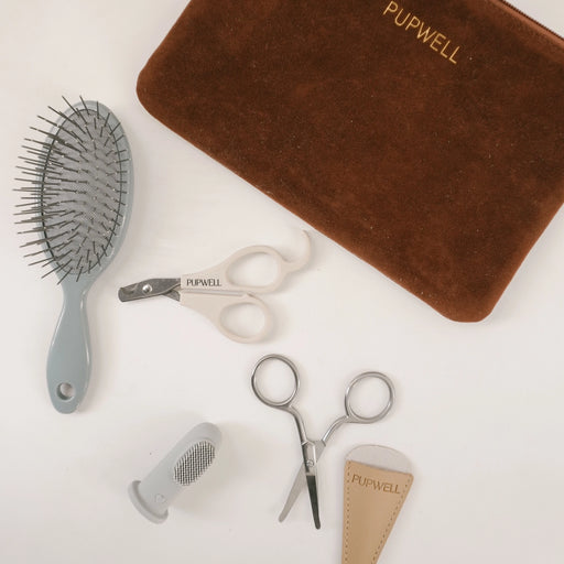 Pupwell grooming kit laid out on a white background, including a gray hairbrush, white nail clippers, silver nail scissors, a gray nail file, and a brown velvet pouch with gold PUPWELL branding.