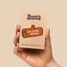 A hand holds a small kraft paper card displaying an enamel pin shaped like a log with the text 'BIG STICK ENERGY' and the brand name 'Scout's Honour' printed above it, against a plain beige background.
