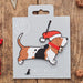 A personalized Christmas beagle ornament on its custom grey backing card, displayed on a rustic wooden surface with holiday decorations.
