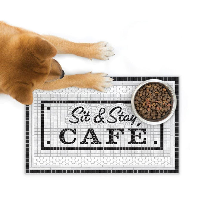 Placemat with small black and white tile motif reading "sit and stay cafe" is pictured on a white background. On top of the placemat a Shiba Inu dog and its food bowl are placed,