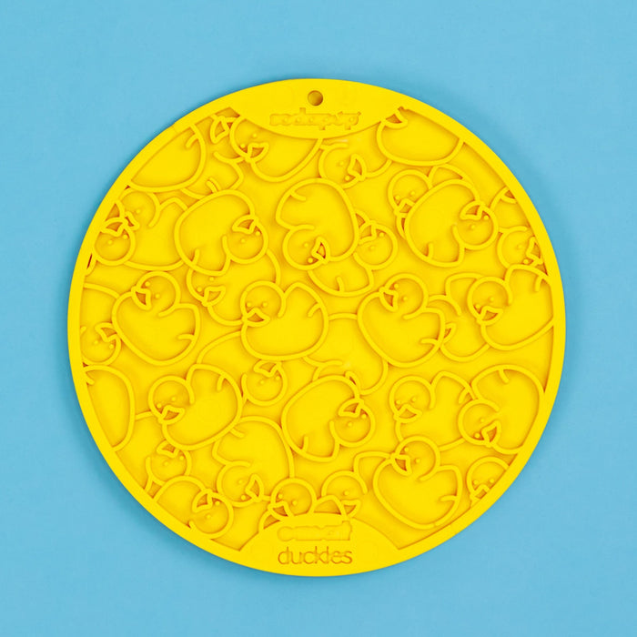 A yellow Duckies Lick Mat for Dogs with a pattern of duck shapes designed for dispensing treats, displayed on a blue background.
