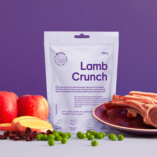 The image shows a bag off Buddy Lamb Crunch dog treats, alongside it a rack of lamb, peas and some apples are scattered.